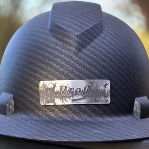May include: A black hard hat with a carbon fiber weave pattern. A silver nameplate displays the name "Brett" with a nature scene of a deer, birds, and water. The hard hat has a brim and side attachments.