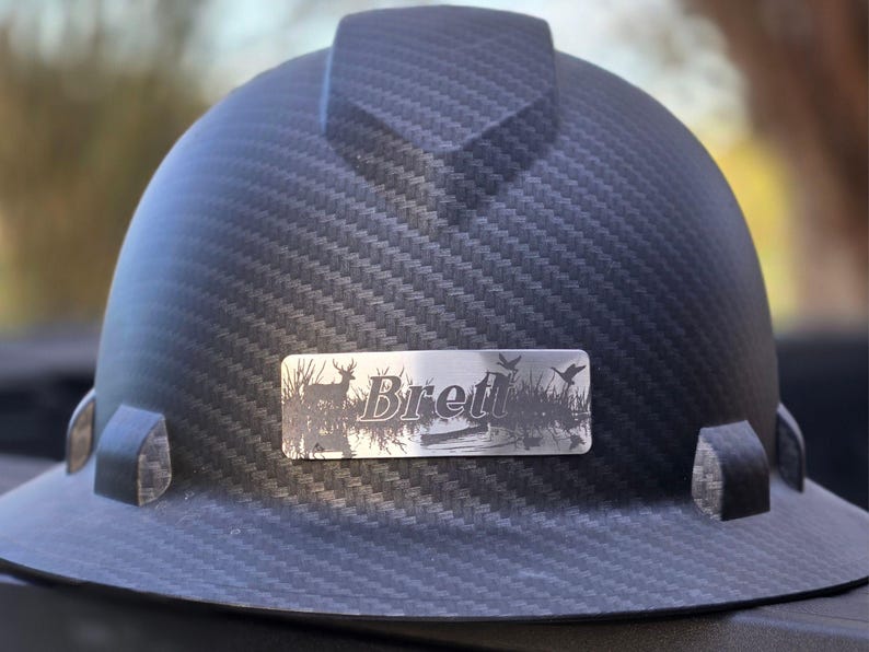 May include: A black hard hat with a carbon fiber weave pattern. A silver nameplate displays the name "Brett" with a nature scene of a deer, birds, and water. The hard hat has a brim and side attachments.