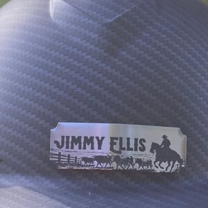 May include: A dark gray hard hat with a carbon fiber texture featuring a rectangular silver metal nameplate. The plate is engraved with the text JIMMY ELLIS above a silhouette graphic of a person riding a horse and a small herd of cattle.