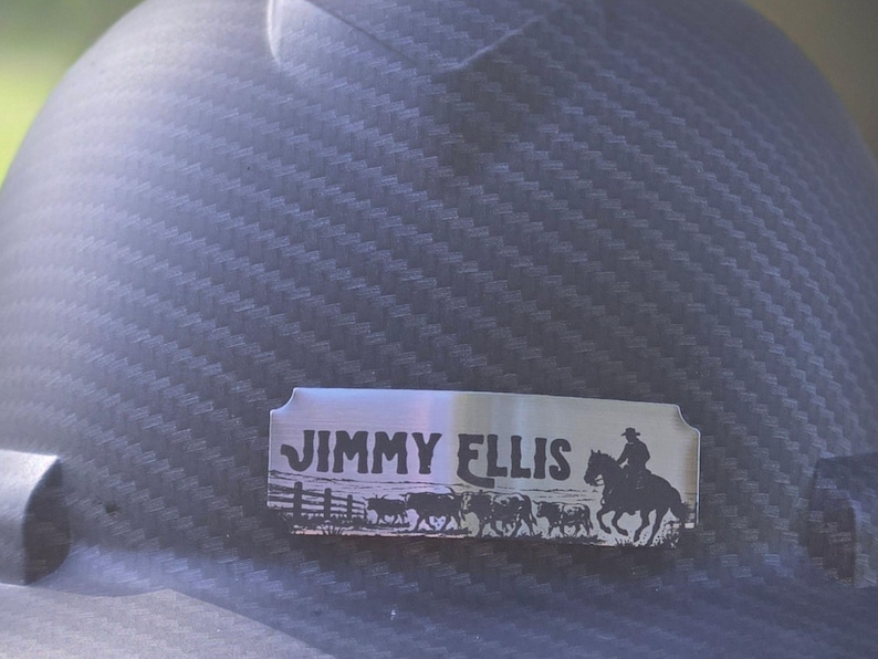 May include: A dark gray hard hat with a carbon fiber texture featuring a rectangular silver metal nameplate. The plate is engraved with the text JIMMY ELLIS above a silhouette graphic of a person riding a horse and a small herd of cattle.