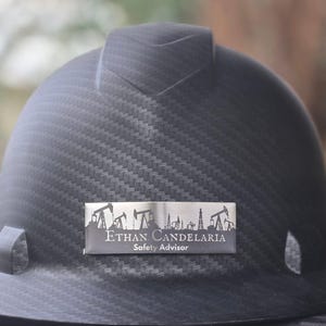 May include: A black hard hat with a textured pattern. A silver nameplate displays the name "Ethan Candelaria" and the title "Safety Advisor" along with an illustration of oil derricks. The hard hat has a curved brim and side attachments.