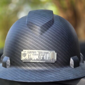 May include: A black hard hat with a carbon fiber weave pattern. A silver nameplate reads "Shonda Williams, Safety Lead" with a city skyline graphic. The hat has a brim and a top ridge.