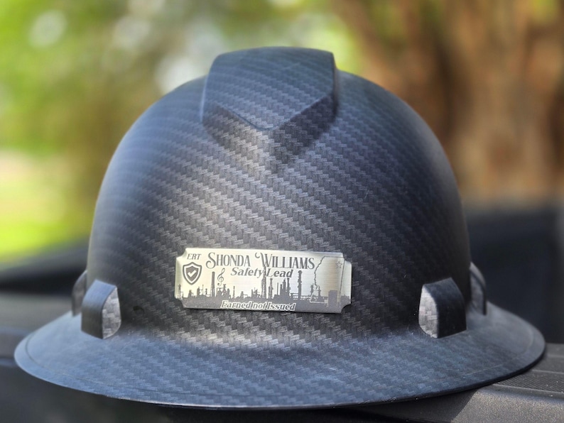 May include: A black hard hat with a carbon fiber weave pattern. A silver nameplate reads "Shonda Williams, Safety Lead" with a city skyline graphic. The hat has a brim and a top ridge.