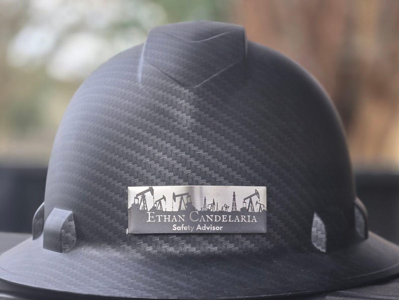 May include: A black hard hat with a textured pattern. A silver nameplate displays the name "Ethan Candelaria" and the title "Safety Advisor" along with an illustration of oil derricks. The hard hat is designed for construction or industrial work.