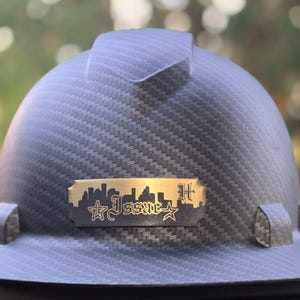 May include: A dark gray hard hat with a textured pattern. A gold-colored metal plate features a city skyline, stars, and the word "Issac" in stylized lettering. The letter "H" is also visible.