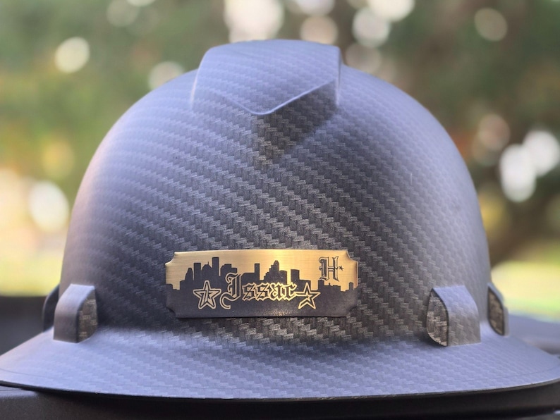 May include: A dark gray hard hat with a textured pattern. A gold-colored metal plate features a city skyline, stars, and the word "Issac" in stylized lettering. The letter "H" is also visible.
