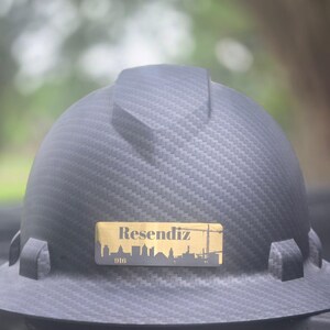 May include: A black carbon fiber hard hat featuring a rectangular gold-colored decal on the front. The decal displays the name Resendiz in a serif font above a silhouette of a city skyline and a construction crane, with the number 916 below.