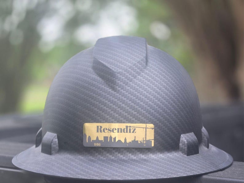 May include: A black carbon fiber hard hat featuring a rectangular gold-colored decal on the front. The decal displays the name Resendiz in a serif font above a silhouette of a city skyline and a construction crane, with the number 916 below.