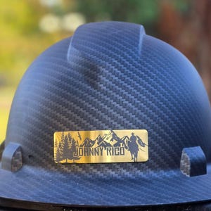 May include: A dark blue hard hat with a textured pattern. A gold-colored rectangular emblem features a mountain range, trees, a cowboy on a horse, and the text "JOHNNY RICO".
