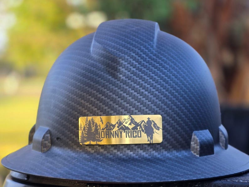 May include: A dark blue hard hat with a textured pattern. A gold-colored rectangular emblem features a mountain range, trees, a cowboy on a horse, and the text "JOHNNY RICO".