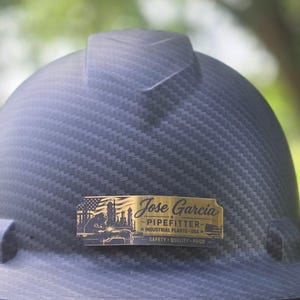 May include: A black carbon fiber textured hard hat featuring a rectangular brass-colored metal plate attached to the front. The plate displays the text Jose Garcia, PIPEFITTER, INDUSTRIAL PLANTS USA, and SAFETY QUALITY PRIDE with a welder graphic.