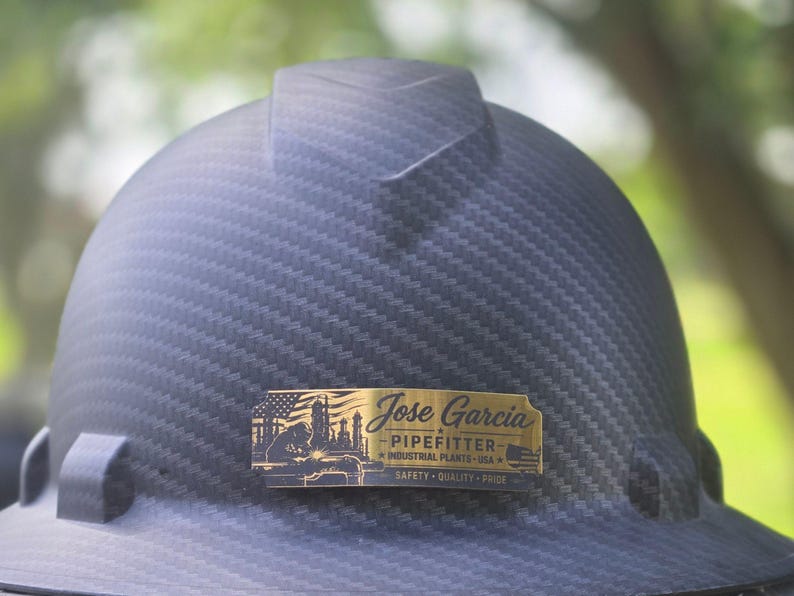 May include: A black carbon fiber textured hard hat featuring a rectangular brass-colored metal plate attached to the front. The plate displays the text Jose Garcia, PIPEFITTER, INDUSTRIAL PLANTS USA, and SAFETY QUALITY PRIDE with a welder graphic.