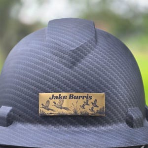 May include: A black carbon fiber pattern hard hat featuring a rectangular gold-toned metal nameplate centered on the front. The nameplate displays the text Jake Burris in black lettering above a graphic of several flying pheasants in a field.