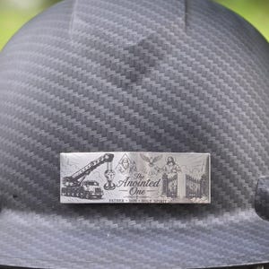 May include: A black carbon fiber hard hat featuring a rectangular metal plate with an engraved design. The plate displays a crane, religious figures, a dove, and the text The Anointed One, Father, Son, Holy Spirit.