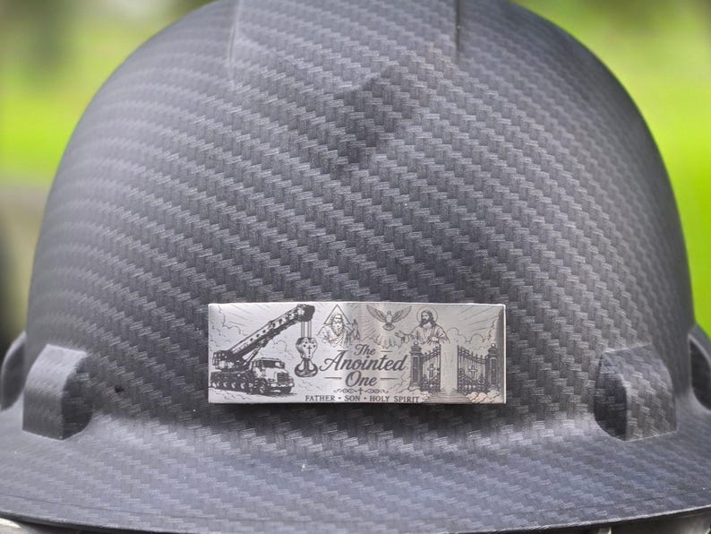 May include: A black carbon fiber hard hat featuring a rectangular metal plate with an engraved design. The plate displays a crane, religious figures, a dove, and the text The Anointed One, Father, Son, Holy Spirit.