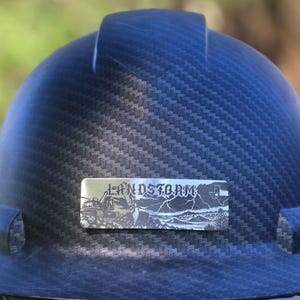May include: A black carbon fiber hard hat featuring a rectangular metal plate on the front. The plate displays the text LANDSTORM above an engraved landscape scene with heavy machinery, lightning, and a small outline of the state of Texas.