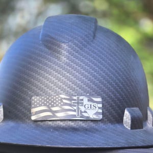May include: A black carbon fiber hard hat featuring a rectangular decal on the front. The decal displays a graphic of the American flag, a Christian cross, the outline of the state of Texas, and the text GIS CONSTRUCTION SERVICES.