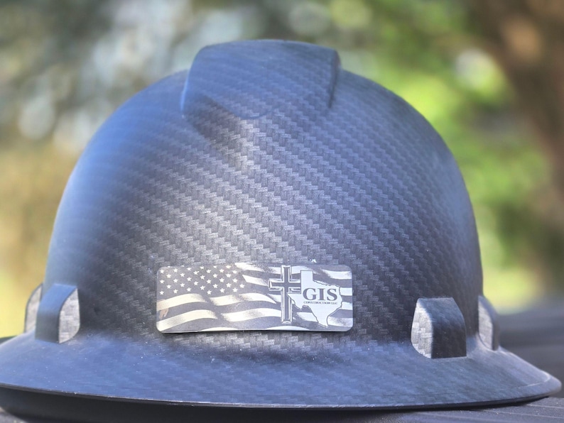 May include: A black carbon fiber hard hat featuring a rectangular decal on the front. The decal displays a graphic of the American flag, a Christian cross, the outline of the state of Texas, and the text GIS CONSTRUCTION SERVICES.