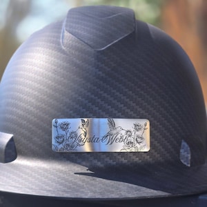 Peut inclure: Casque de chantier noir avec une surface texturée. Une plaque signalétique argentée présente un motif de colibri et de roses avec le nom "Krysta Webb" gravé. Le casque est conçu pour la construction ou l'usage industriel.