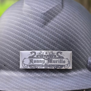 May include: A dark gray hard hat with a carbon fiber texture pattern. A rectangular silver nameplate is attached to the front, featuring a black graphic design of a skull and two snakes, with the text Ronny Murillo printed in the center.