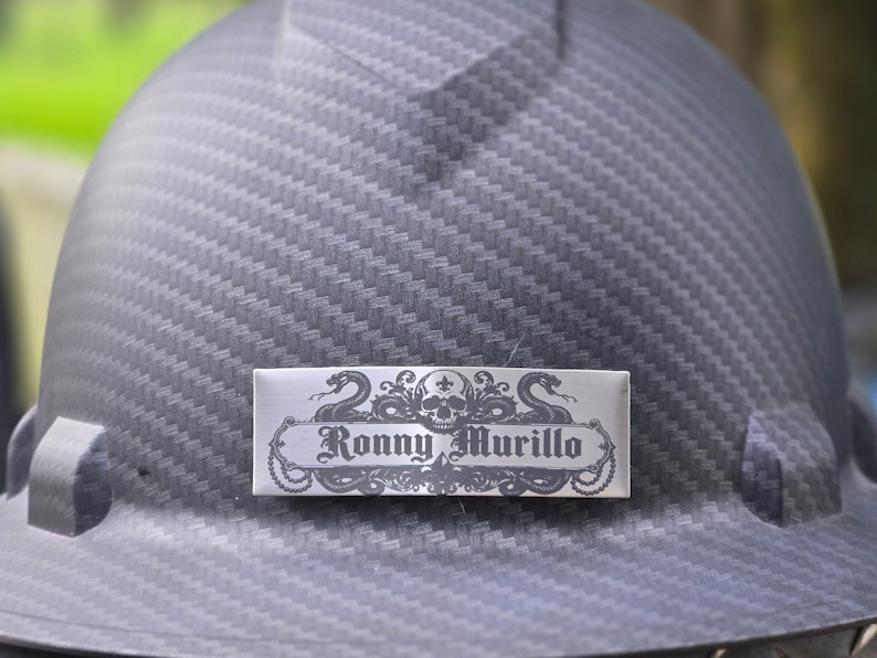 May include: A dark gray hard hat with a carbon fiber texture pattern. A rectangular silver nameplate is attached to the front, featuring a black graphic design of a skull and two snakes, with the text Ronny Murillo printed in the center.