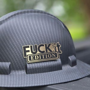 May include: A black hard hat with a textured surface. A gold-colored emblem with the words "FUCK IT EDITION" is affixed to the front of the hat. The hat is designed for construction or industrial use.