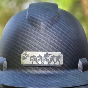 May include: Black hard hat with a textured pattern. A silver nameplate displays the word "BIGGEN" and silhouettes of soldiers. The hard hat has a brim and a top band.
