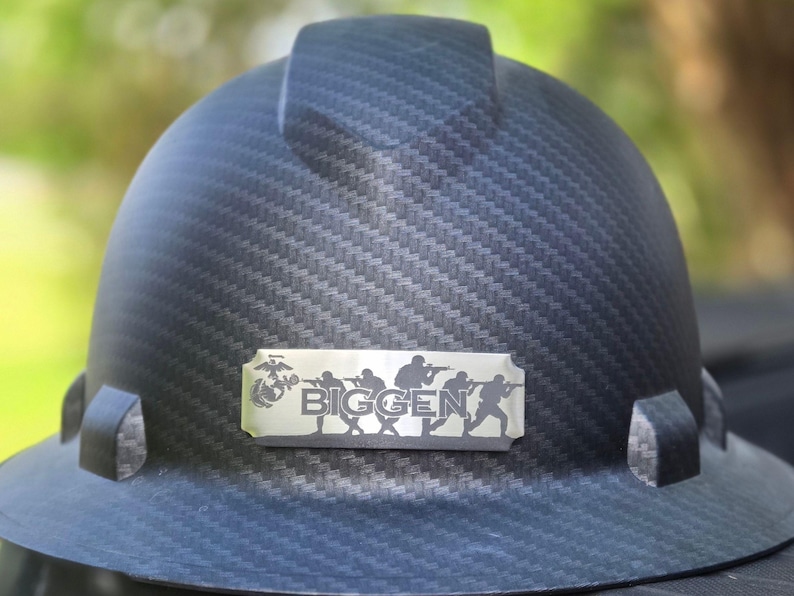 May include: Black hard hat with a textured pattern. A silver nameplate displays the word "BIGGEN" and silhouettes of soldiers. The hard hat has a brim and a top band.