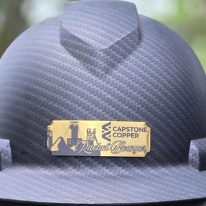 May include: A dark gray hard hat with a textured pattern. A gold-colored nameplate displays the text "Capstone Copper" and "Rachel Bowyer" along with a silhouette of a mining scene. The hard hat is designed for construction or industrial use.