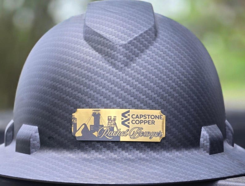 May include: A dark gray hard hat with a textured pattern. A gold-colored nameplate displays the text "Capstone Copper" and "Rachel Bowyer" along with a silhouette of a mining scene. The hard hat is designed for construction or industrial use.