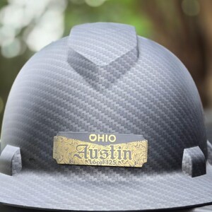 May include: A gray hard hat with a carbon fiber pattern. A gold-colored nameplate reads "OHIO Austin Local 125". The hard hat has a brim and a top ridge.
