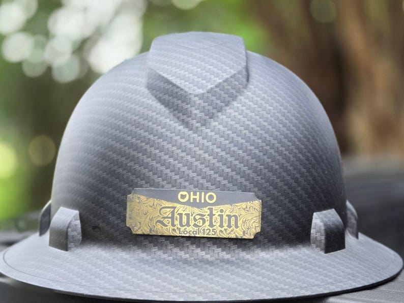 May include: A gray hard hat with a carbon fiber pattern. A gold-colored nameplate reads "OHIO Austin Local 125". The hard hat has a brim and a top ridge.