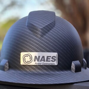 May include: A black hard hat with a carbon fiber weave pattern. A silver nameplate displays the text "NAES Power Contractors" and a logo. The hard hat has a brim and side attachments.
