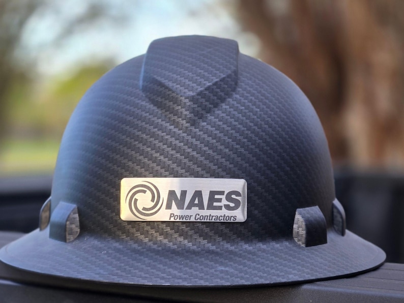 May include: A black hard hat with a carbon fiber weave pattern. A silver nameplate displays the text "NAES Power Contractors" and a logo. The hard hat has a brim and side attachments.