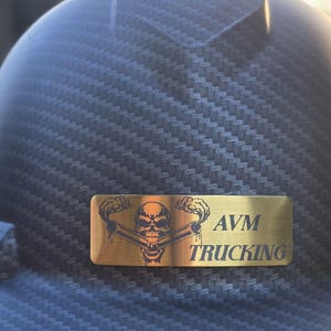 May include: Close-up of a dark gray hard hat with a carbon fiber weave pattern. A gold-colored rectangular emblem features a skull with crossed pipes and the text "AVM TRUCKING".