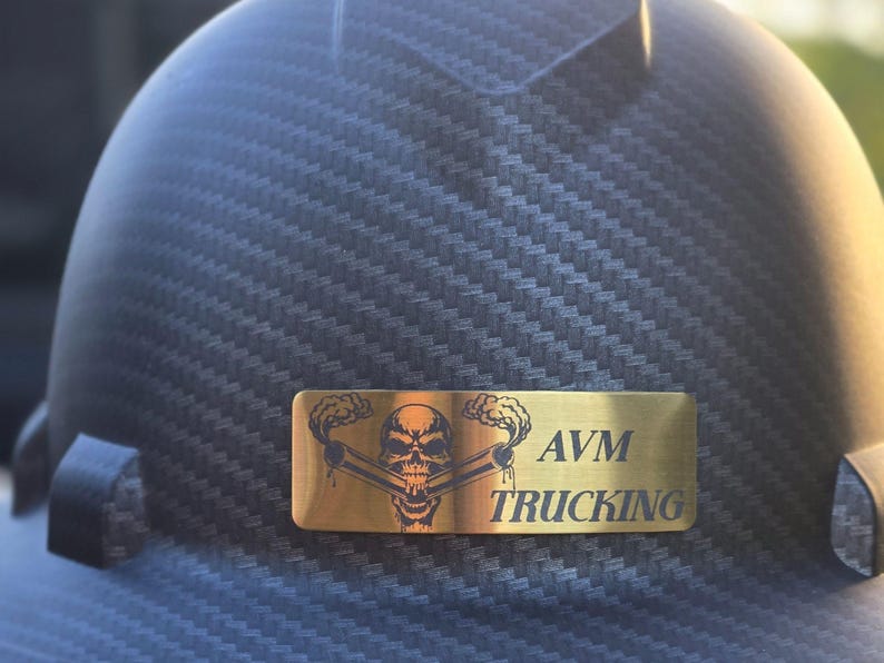 May include: Close-up of a dark gray hard hat with a carbon fiber weave pattern. A gold-colored rectangular emblem features a skull with crossed pipes and the text "AVM TRUCKING".