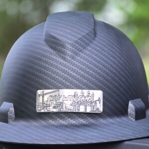 May include: A black carbon fiber hard hat featuring a central ridge and a rectangular silver-toned metal plate on the front. The plate displays an engraved illustration of a construction site with a truck and the text KYLE WILLIAMS.
