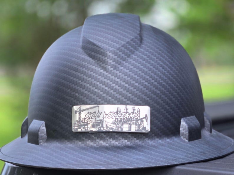 May include: A black carbon fiber hard hat featuring a central ridge and a rectangular silver-toned metal plate on the front. The plate displays an engraved illustration of a construction site with a truck and the text KYLE WILLIAMS.