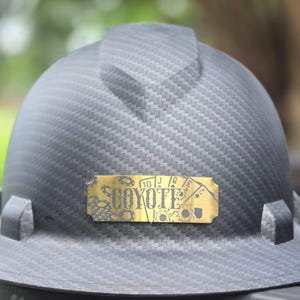 May include: A dark gray hard hat with a textured pattern. A gold-colored nameplate features the word "COYOTE" and playing card symbols. The hard hat is designed for construction or industrial use.