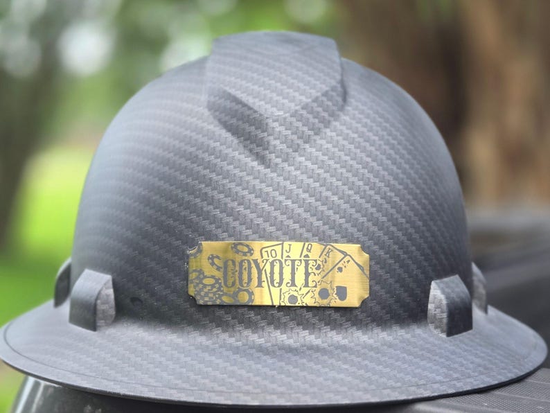 May include: A dark gray hard hat with a textured pattern. A gold-colored nameplate features the word "COYOTE" and playing card symbols. The hard hat is designed for construction or industrial use.