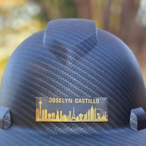 May include: A black hard hat with a textured surface. A gold nameplate displays the name "JOSELYN CASTILLO" above a skyline silhouette. The skyline includes the Eiffel Tower, Statue of Liberty, and other landmarks.