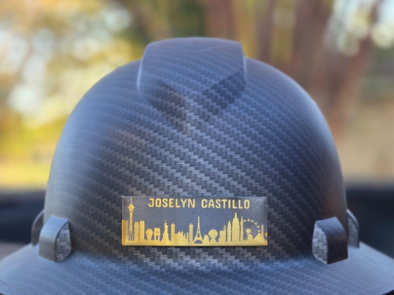 May include: A black hard hat with a textured surface. A gold nameplate displays the name "JOSELYN CASTILLO" above a skyline silhouette. The skyline includes the Eiffel Tower, Statue of Liberty, and other landmarks.