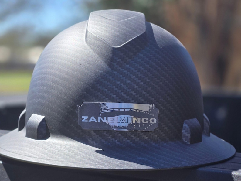 May include: A dark gray hard hat with a textured surface. The hard hat has a rectangular label with the text "ZANE MINGO" and a bridge graphic. The hat is designed for construction or industrial use.