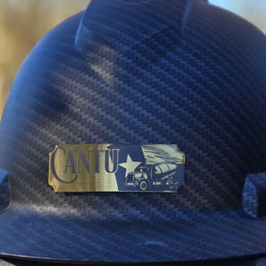 May include: A black hard hat with a carbon fiber texture pattern. Attached to the front is a rectangular gold-toned metal plate featuring the text CANTU, a five-pointed star, and a graphic illustration of a cement mixer truck in black.