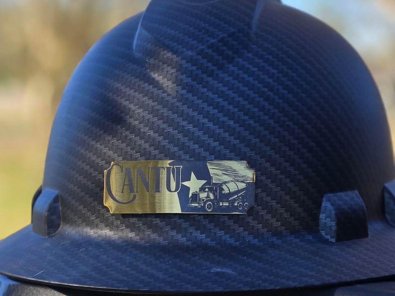 May include: A black hard hat with a carbon fiber texture pattern. Attached to the front is a rectangular gold-toned metal plate featuring the text CANTU, a five-pointed star, and a graphic illustration of a cement mixer truck in black.
