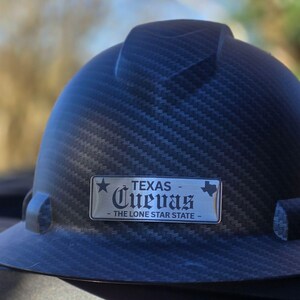 May include: A dark gray hard hat with a textured pattern. A silver nameplate displays the word "TEXAS" above "Cuevas" and "THE LONE STAR STATE". The hard hat is designed for construction or industrial use.
