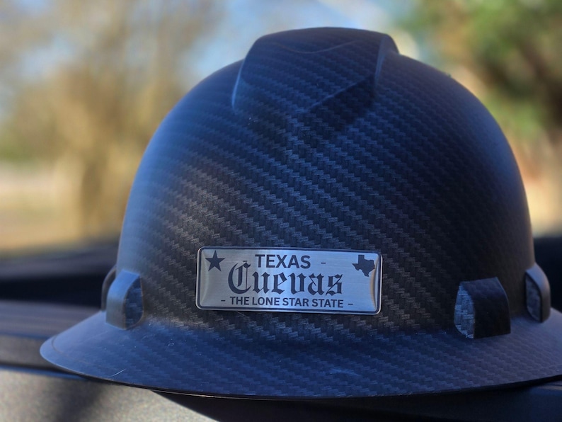 May include: A dark gray hard hat with a textured pattern. A silver nameplate displays the word "TEXAS" above "Cuevas" and "THE LONE STAR STATE". The hard hat is designed for construction or industrial use.