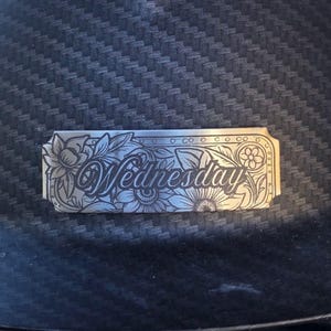 May include: A silver-colored metal plate with the word "Wednesday" engraved in an elegant script, surrounded by floral and leaf designs. The plate is set against a dark, textured background.