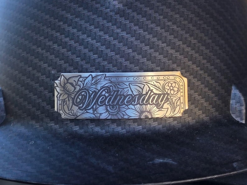May include: A silver-colored metal plate with the word "Wednesday" engraved in an elegant script, surrounded by floral and leaf designs. The plate is set against a dark, textured background.