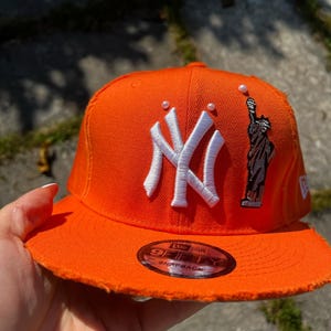 Op de afbeelding: Een oranje snapback-pet met een wit geborduurd New York Yankees-logo, een grijze patch van het Vrijheidsbeeld en kleine witte parelaccenten. De klep heeft een gerafelde rand en een ronde sticker met de tekst 9FIFTY SNAPBACK.